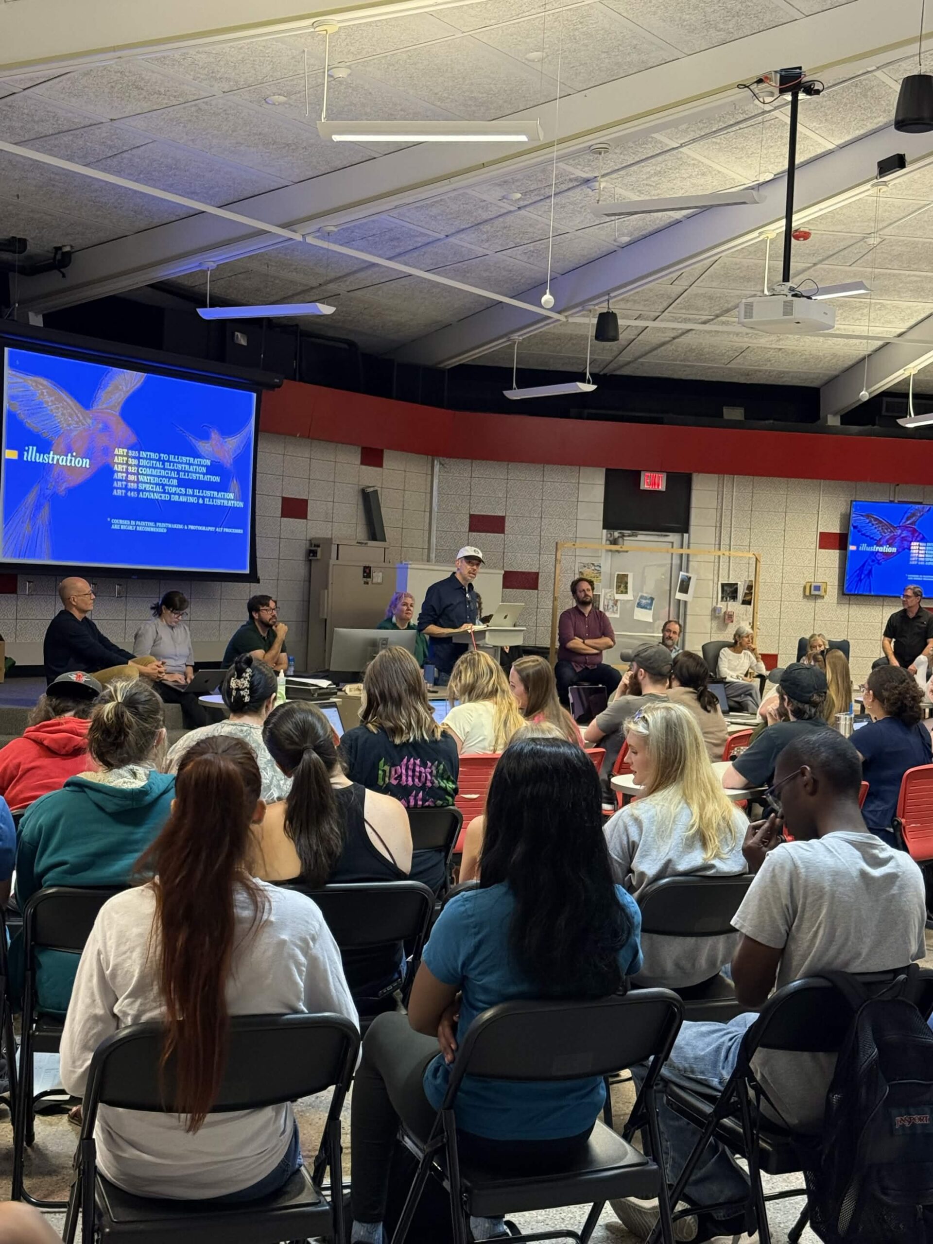 A large group of students sits in a classroom listening to a faculty panel at the front, with a screen displaying a slide about the illustration program.