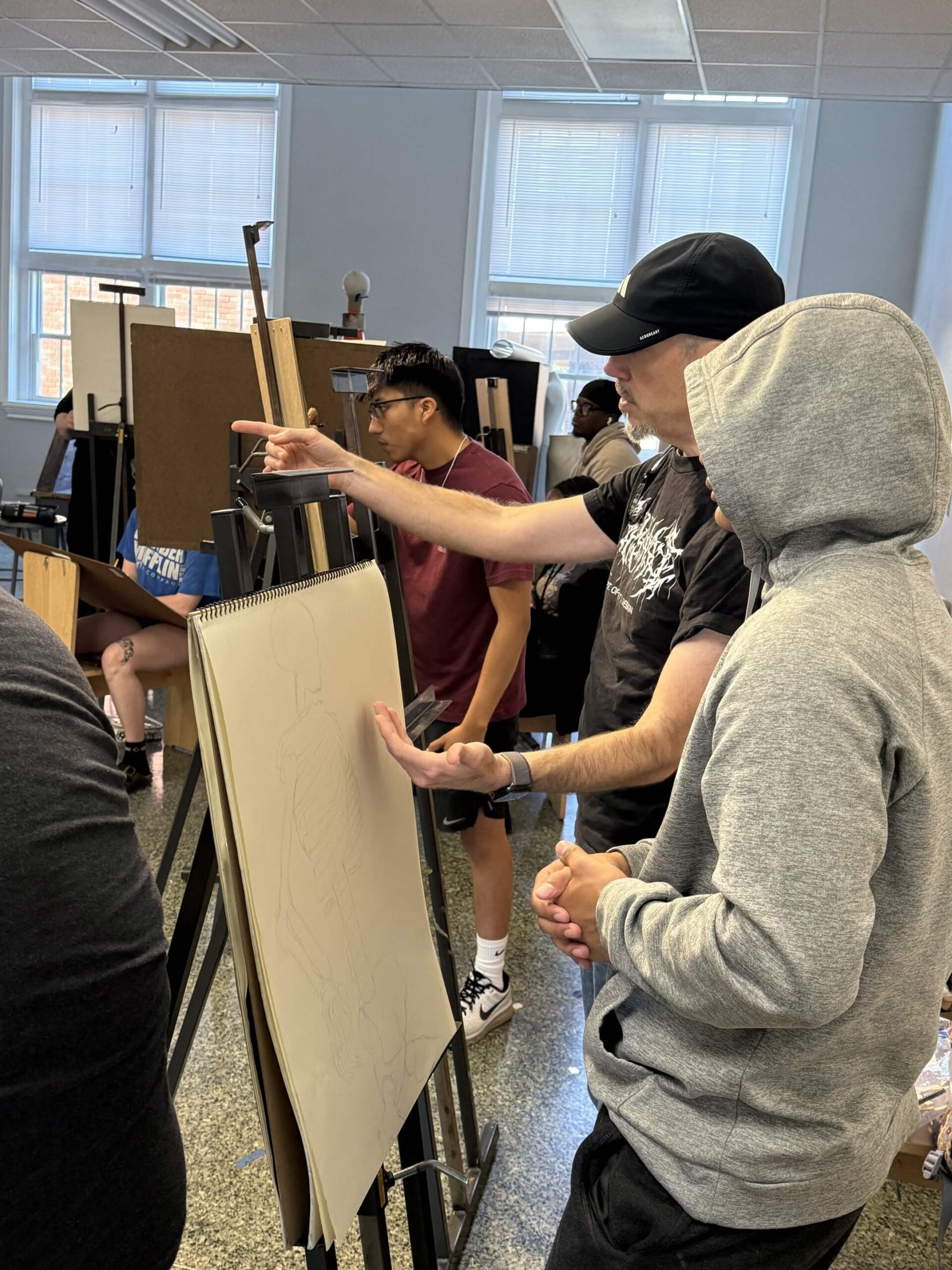 An instructor points at a large charcoal figure drawing on an easel while students gather around in the figure drawing studio.