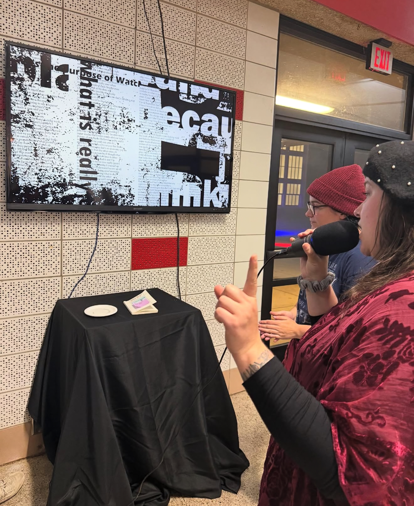 At a gallery opening, a speaker gestures while talking into a microphone near a screen displaying fragmented black and white text artwork and a covered pedestal.