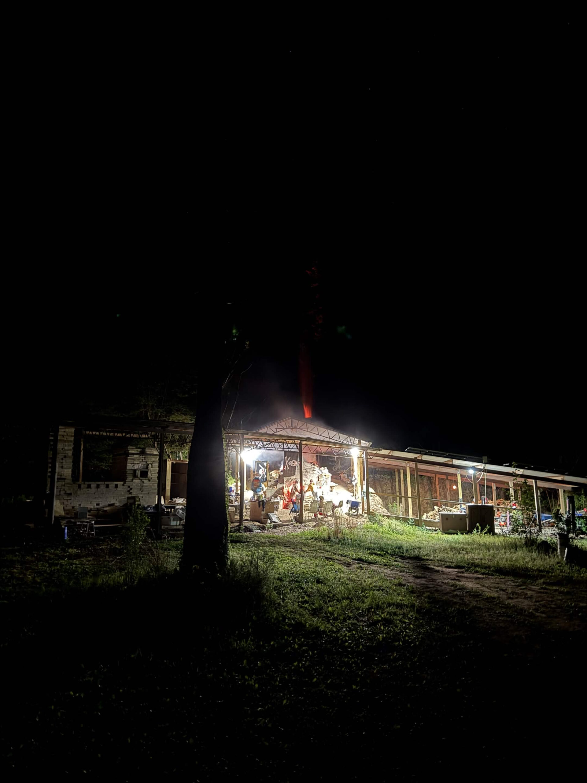 Nighttime photo of a glowing wood fired kiln with flames visible inside its open structure, with a tree silhouetted in the foreground.