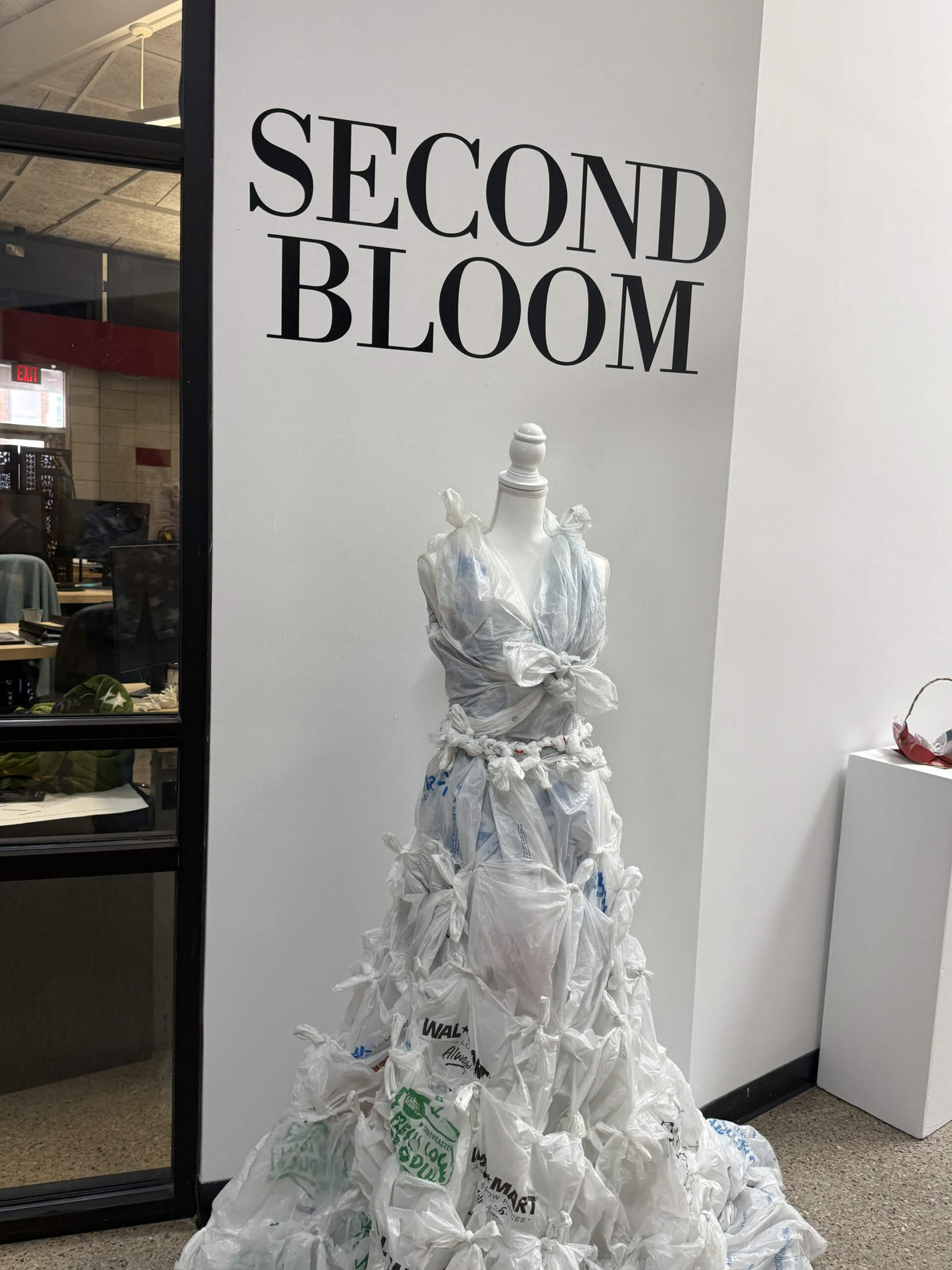 Gallery display titled "Second Bloom," showing a mannequin dressed in a gown made entirely of plastic shopping bags, including a Walmart bag.