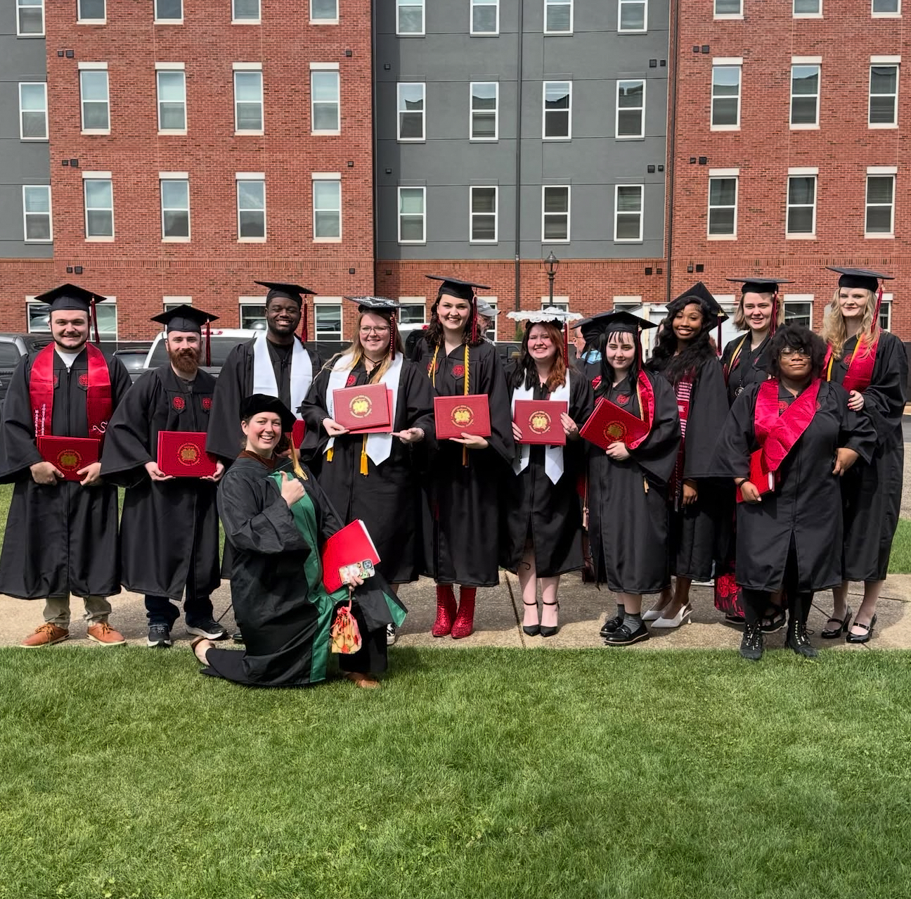 Group of graduates in caps and gowns holding red diploma covers, posed on a lawn outside a brick residence hall, with a faculty member kneeling in front in academic regalia.