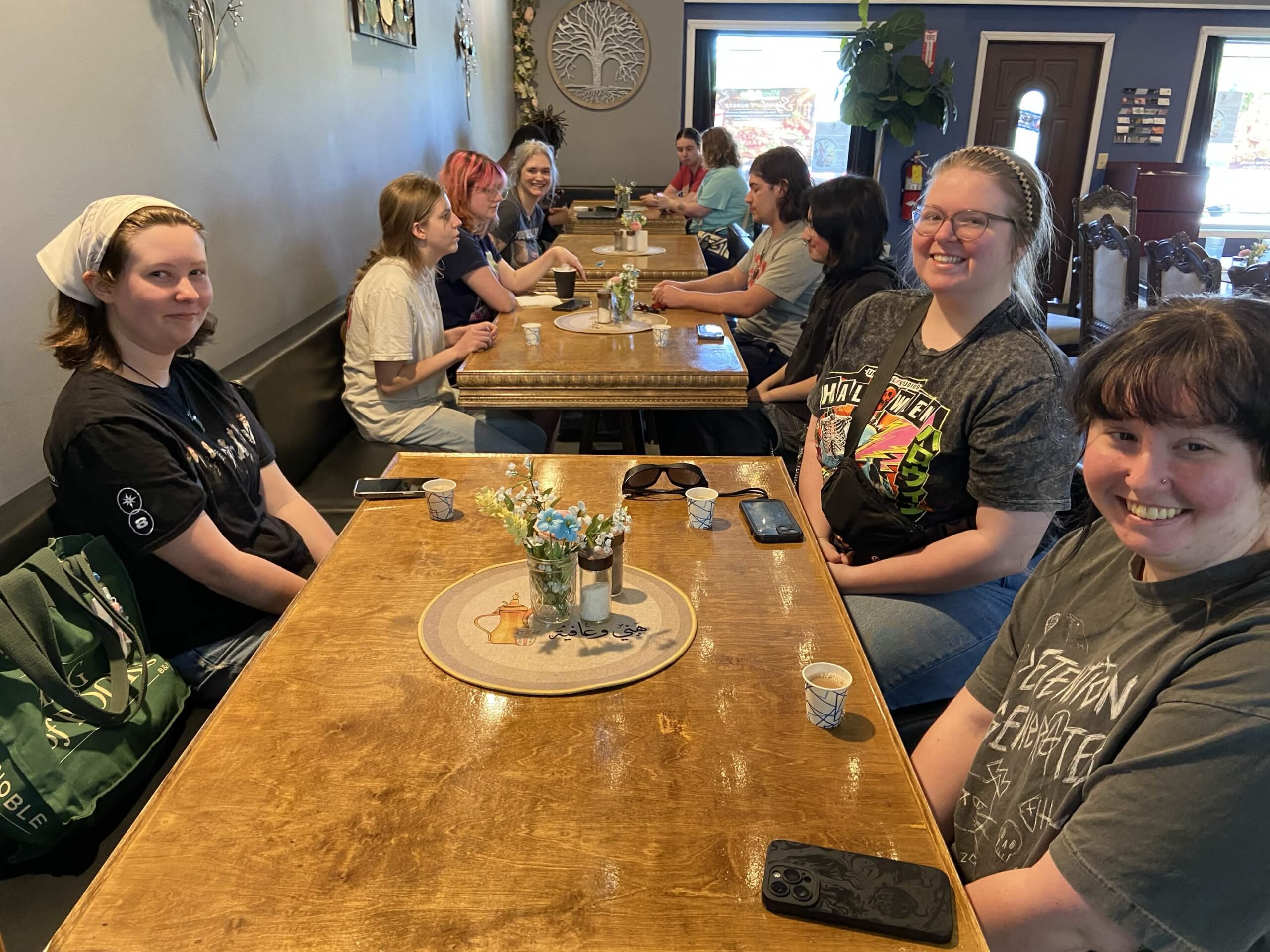 Group of women smiling and seated together at wooden tables in a restaurant, with drinks and phones on the tables.