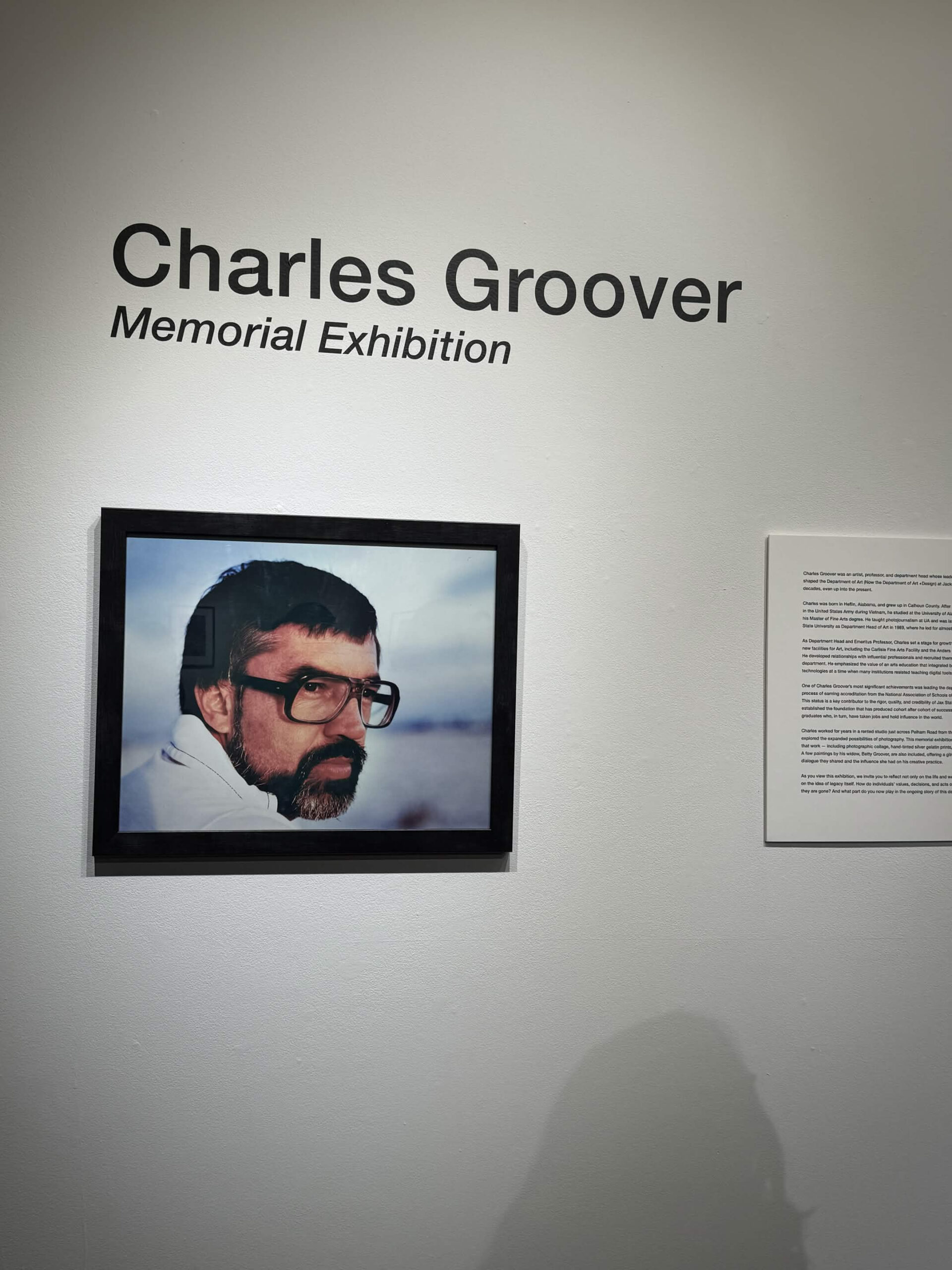 Gallery wall reading "Charles Groover Memorial Exhibition" beside a framed portrait photo of a man with glasses and a beard and a text panel.
