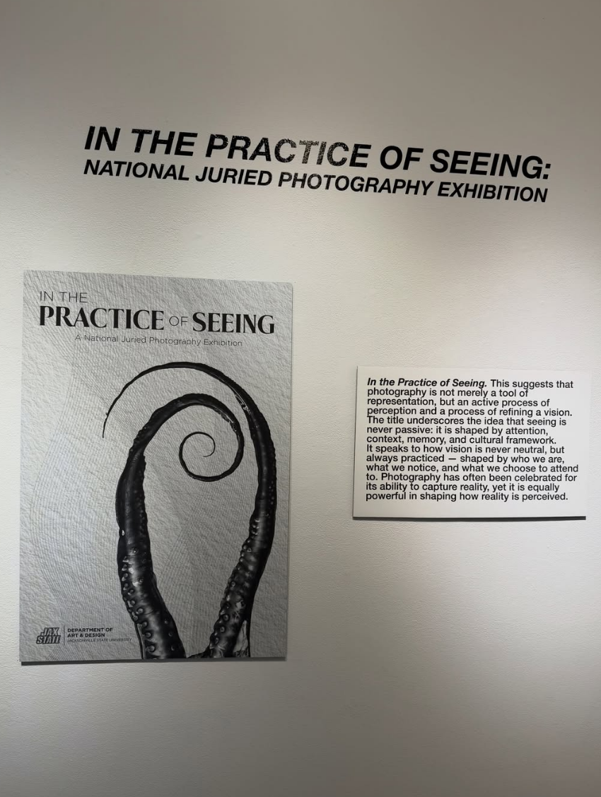 Gallery wall display for "In the Practice of Seeing: A National Juried Photography Exhibition," with a poster showing a spiraled black and white tentacle photograph and an artist statement placard.